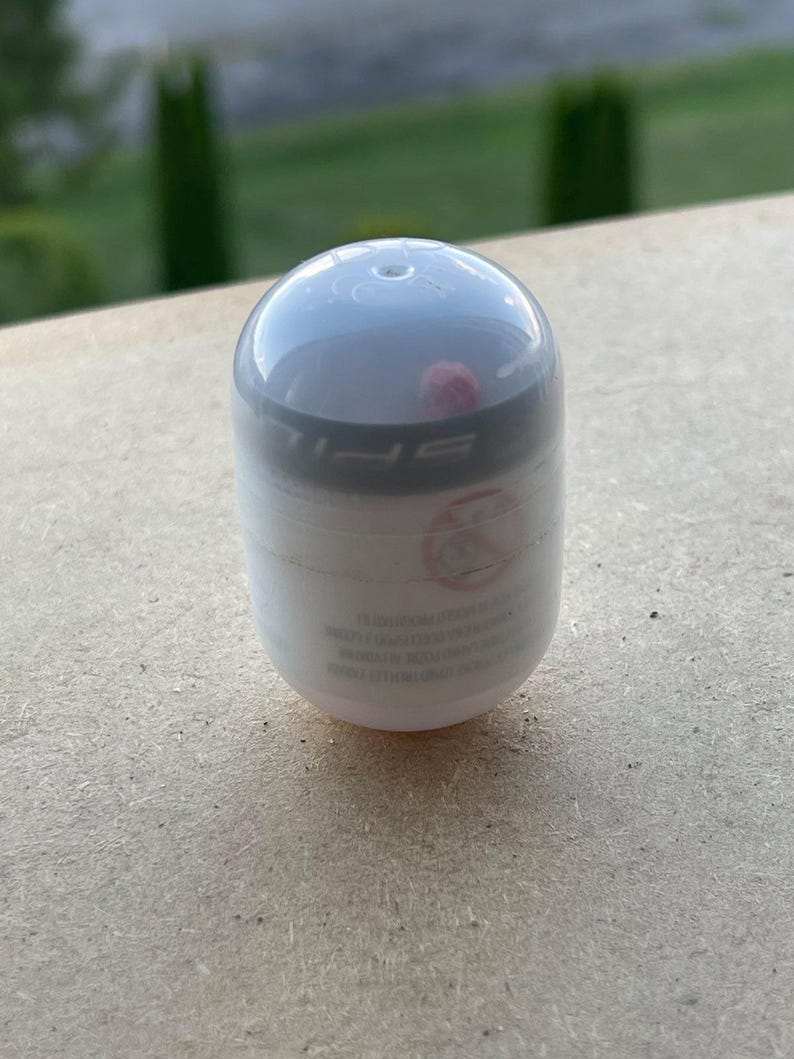 Pode incluir: Um recipiente branco e cinza em forma de c&aacute;psula com uma parte superior arredondada. O recipiente tem uma faixa preta ao redor do meio e algum texto. O fundo &eacute; uma superf&iacute;cie marrom clara e folhagem verde.