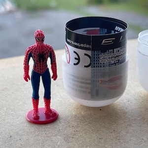 Pode incluir: Uma figura do Homem-Aranha, vermelha e azul, com um emblema de aranha preta, est&aacute; em p&eacute; sobre uma base vermelha. Ao lado, uma c&aacute;psula parcialmente aberta, branca e transparente, com um r&oacute;tulo em preto e branco.