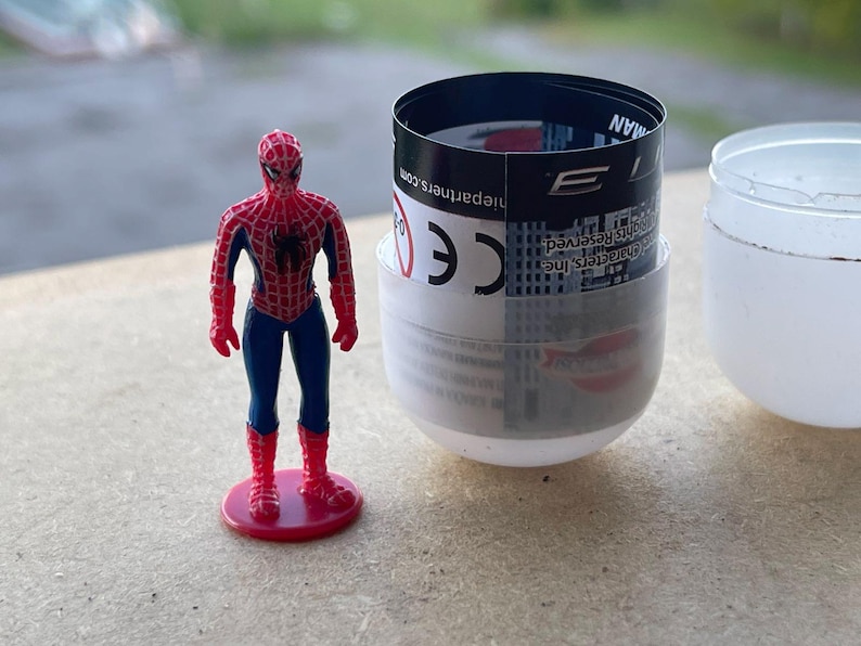 Pode incluir: Uma figura do Homem-Aranha, vermelha e azul, com um emblema de aranha preta, est&aacute; em p&eacute; sobre uma base vermelha. Ao lado, uma c&aacute;psula parcialmente aberta, branca e transparente, com um r&oacute;tulo em preto e branco.