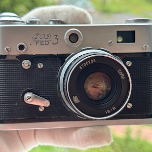 May include: A silver and black FED 3 rangefinder camera featuring a textured black body, a silver top plate with the text FED 3 in Cyrillic and English, and a black lens with the markings 8708238, 2,8/52, and M-61 printed on the front ring.