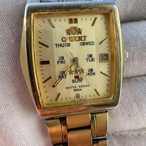 May include: A gold-toned Orient wristwatch with a rectangular face. The watch face is gold with black hands and markers, displaying the day, date, and time. The watch band is also gold-toned. The watch reads "WATER RESIST 50M".