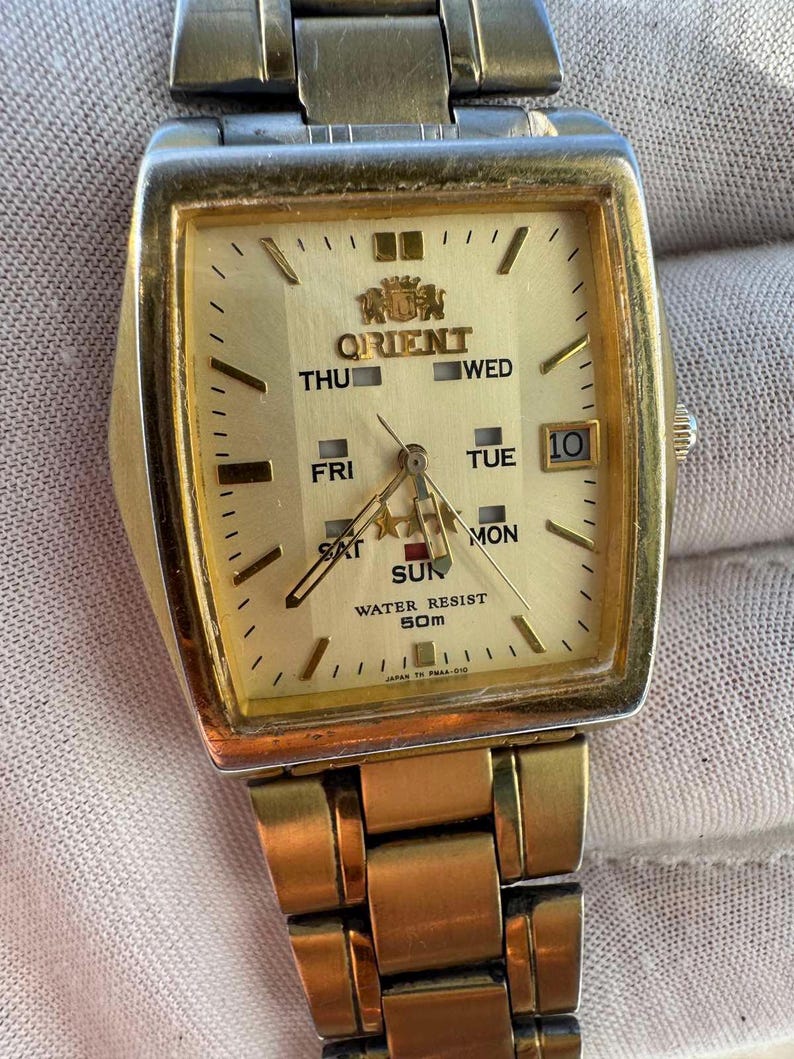 May include: A gold-toned Orient wristwatch with a rectangular face. The watch face is gold with black hands and markers, displaying the day, date, and time. The watch band is also gold-toned. The watch reads "WATER RESIST 50M".