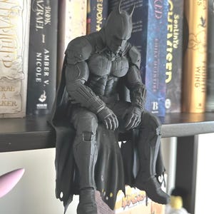 May include: A matte dark grey Batman figurine in a seated position with a downward-facing head and a flowing cape. The character wears a detailed armoured suit with gauntlets and boots, positioned with hands resting on knees, showcasing a textured surface.