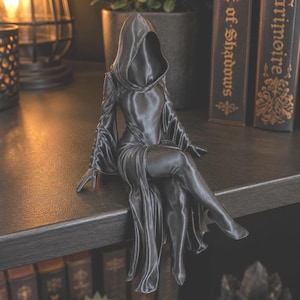 Könnte beinhalten: Eine dunkelgraue, glatte Figur einer sitzenden Gestalt, die einen Kapuzenumhang mit langen, fließenden Ärmeln trägt. Die Figur ist in einer entspannten, sitzenden Pose mit übereinandergeschlagenen Beinen dargestellt und hat eine tiefe Kapuze.