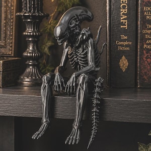 Puede incluir: Una figura detallada de color gris oscuro de una criatura Xenomorfo de la franquicia Alien, representada en posición sentada con las piernas colgando sobre el borde de un estante. La figura presenta una cabeza lisa y alargada, costillas y cola.