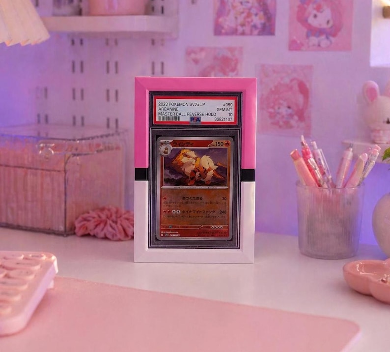 May include: A graded Pok&eacute;mon card in a pink and white holder. The card features an Arcanine illustration and text that reads "2023 POKEMON SV2a JP ARCANINE MASTER BALL REVERSE HOLD". The holder is displayed on a desk with other pink accessories.