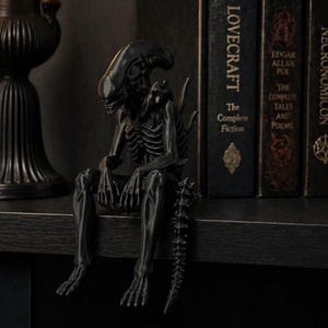Puede incluir: Una figura negra de Alien se sienta en una estantería junto a libros con títulos como "Lovecraft" y "Necronomicon". La figura tiene un diseño esquelético y una larga cola. La estética general es oscura y gótica.