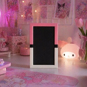 May include: A pink and white framed chalkboard sits on a desk, surrounded by pink decor. A neon pink bow-shaped light, a keyboard, and a mouse are visible. A vase of pink tulips and a bunny-shaped lamp add to the aesthetic.