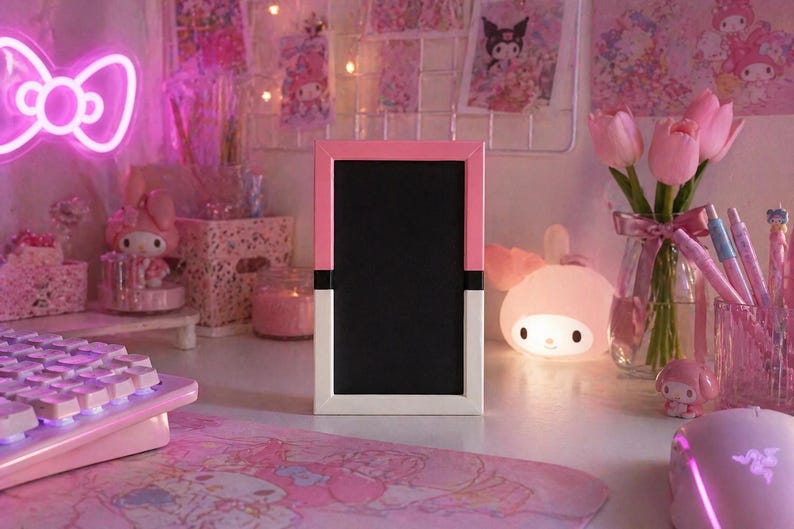 May include: A pink and white framed chalkboard sits on a desk, surrounded by pink decor. A neon pink bow-shaped light, a keyboard, and a mouse are visible. A vase of pink tulips and a bunny-shaped lamp add to the aesthetic.
