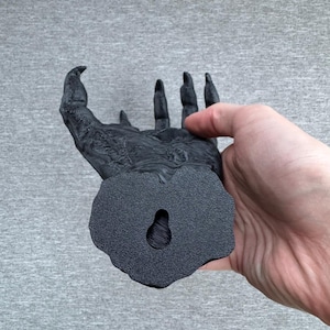 Puede incluir: Una escultura de pared negra y texturizada con forma de mano con garras y dedos largos y afilados. La base presenta un orificio en forma de l&aacute;grima para colgar, y la superficie muestra detalles intrincados y rugosos que recuerdan a piel.