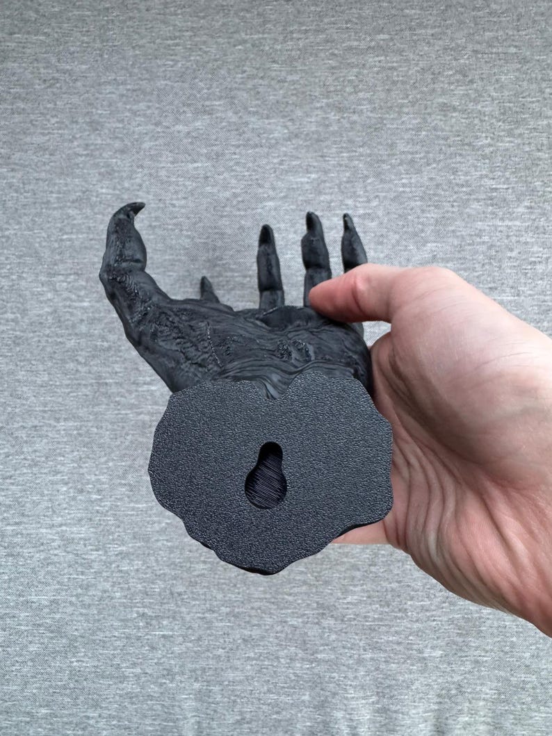 Puede incluir: Una escultura de pared negra y texturizada con forma de mano con garras y dedos largos y afilados. La base presenta un orificio en forma de l&aacute;grima para colgar, y la superficie muestra detalles intrincados y rugosos que recuerdan a piel.