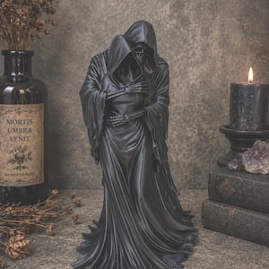 May include: A black sculpture depicting two hooded skeletal figures in a close embrace. The figures are draped in long, flowing robes that pool at the base. The skeletal hands are visible, resting on the back and waist of the other figure.
