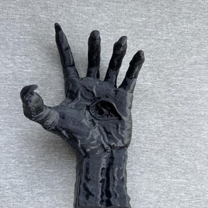 Puede incluir: Una escultura negra y texturizada de una mano con dedos alargados en forma de garra y un ojo &uacute;nico y prominente incrustado en el centro de la palma. La superficie de la mano presenta relieves detallados similares a venas y un acabado brillante.