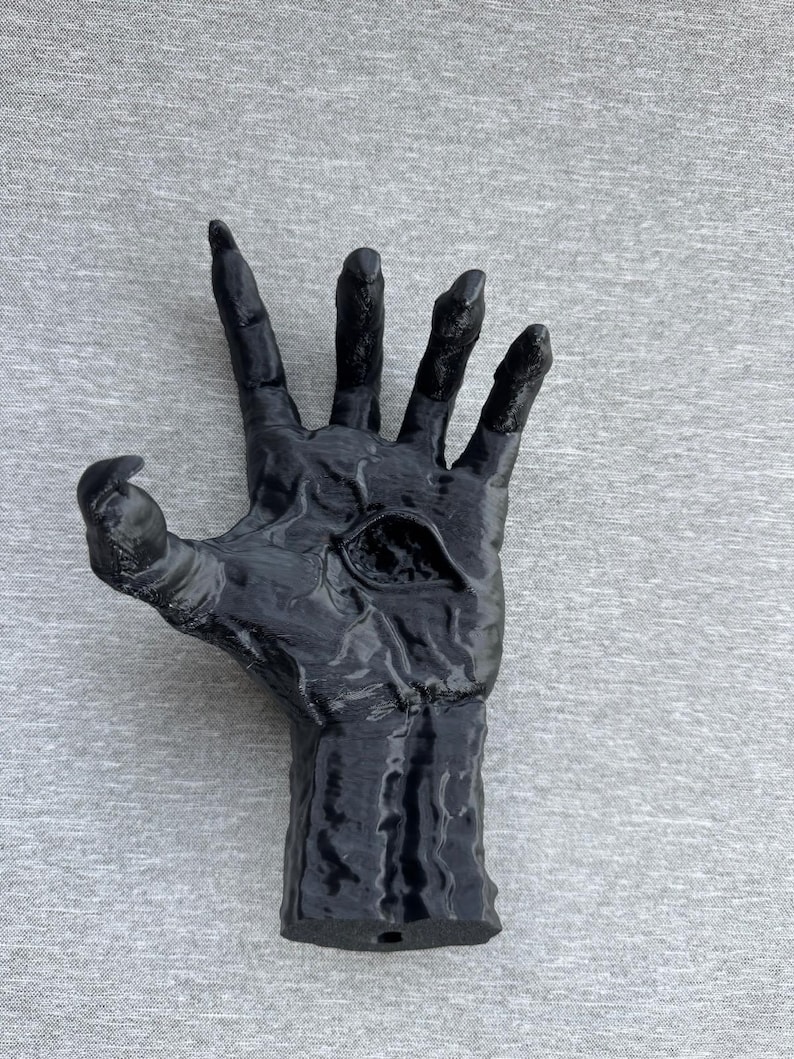 Puede incluir: Una escultura negra y texturizada de una mano con dedos alargados en forma de garra y un ojo &uacute;nico y prominente incrustado en el centro de la palma. La superficie de la mano presenta relieves detallados similares a venas y un acabado brillante.
