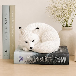 May include: A white fox figurine with black-tipped ears and a black nose, curled into a sleeping position. The fox is resting on top of a black book titled Quicksilver by Callie Hart, which features a small purple butterfly graphic on the cover.