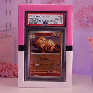 May include: A framed Pok&eacute;mon card featuring Arcanine. The card is encased in a clear plastic holder and set within a pink and white frame. The card's details include Japanese text, a Master Ball Reverse Holo, and a Gem Mint 10 grade.
