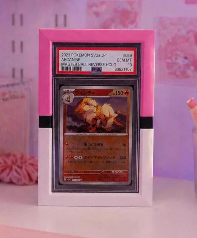 May include: A framed Pok&eacute;mon card featuring Arcanine. The card is encased in a clear plastic holder and set within a pink and white frame. The card's details include Japanese text, a Master Ball Reverse Holo, and a Gem Mint 10 grade.