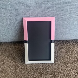 May include: A rectangular photo frame with a black interior. The frame is pink at the top, white at the bottom, and has a black accent in the middle. The frame is sitting on a brown carpet.