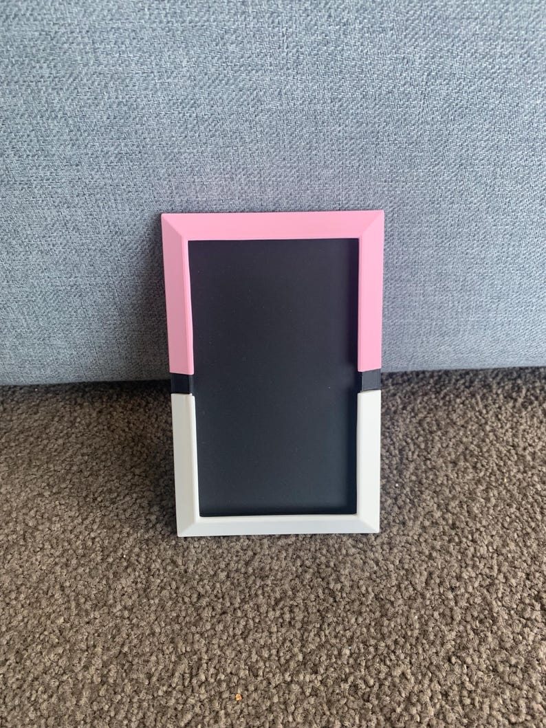 May include: A rectangular photo frame with a black interior. The frame is pink at the top, white at the bottom, and has a black accent in the middle. The frame is sitting on a brown carpet.