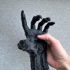 Puede incluir: Una escultura negra impresa en tres dimensiones de una mano con garras y dedos alargados y puntiagudos. La superficie tiene una apariencia texturizada y estratificada caracter&iacute;stica de la fabricaci&oacute;n aditiva, mostrando una forma oscura y r&iacute;gida.