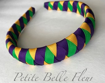May include: A braided headband in purple, gold, and green. The headband is made of woven ribbons in a repeating pattern. The text "Petite Belle Fleur" is printed in a cursive font at the bottom of the image.