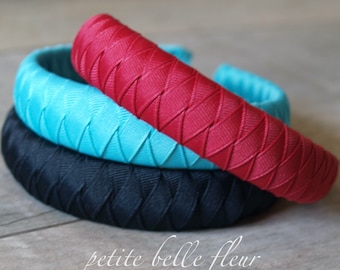 May include: Three braided headbands in black, turquoise, and red. The headbands are made of a woven fabric and are stacked on top of each other.