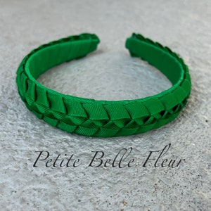 May include: A bright green woven bracelet with an open design. The bracelet is made of a series of woven ribbons, creating a textured surface. The text "Petite Belle Fleur" is printed in black cursive font.