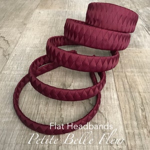 May include: A collection of burgundy flat headbands with diamond-patterned and braided designs. The headbands are arranged on a wooden surface. The text "Flat Headbands" and "Petite Belle Fleur" are visible.