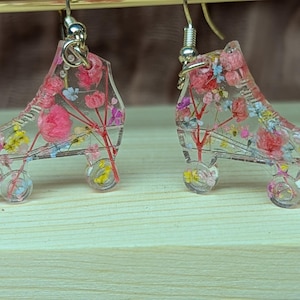 May include: A pair of clear roller skate earrings filled with colorful dried flowers. The earrings have silver-colored hooks and wheels. The flowers are pink, yellow, and blue. The earrings are on a light brown wooden surface.