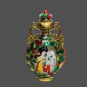 Può includere: Flacone di profumo dorato e ornato con una scena dipinta di una donna in abiti tradizionali. La bottiglia presenta un design floreale verde e rosso, una parte superiore gioiellata e due manici. L'opera d'arte è incorniciata al centro del flacone.
