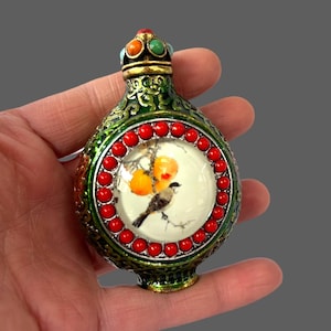 Peut inclure: Une bouteille ornée avec un design détaillé. La bouteille présente une image circulaire centrale d'un oiseau perché sur une branche avec des fruits oranges. La bouteille est verte avec des accents rouges et dorés et un dessus orné de bijoux.