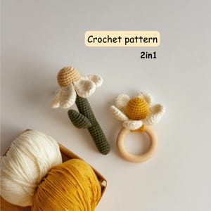 Könnte beinhalten: Zwei gehäkelte Gänseblümchen-Spielzeuge, eines mit einem grünen Stiel und das andere an einem Holzring befestigt. Die Gänseblümchen haben weiße Blütenblätter und gelbe Zentren. Der Text "Crochet pattern 2in1" befindet sich in einer gelben Box. Ein Korb mit Garn befindet sich unten links.
