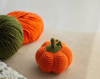 May include: A small, round, orange crocheted pumpkin with a green stem and textured ridges. Behind it sit two large balls of yarn, one in olive green and one in bright orange, alongside a corner of paper featuring a botanical leaf pattern.