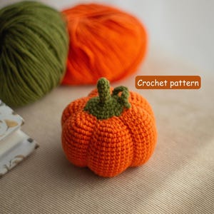 Puede incluir: Una calabaza naranja tejida a ganchillo con un tallo verde. La imagen también muestra dos ovillos de lana, uno naranja y otro verde. El texto "Crochet pattern" está en un rectángulo marrón.