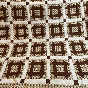 May include: A brown and white quilt with a geometric pattern. The quilt is made up of squares with a brown center and white borders. The squares are arranged in a grid pattern.