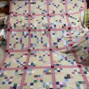 May include: A large patchwork quilt with a cream background and a pink border. The quilt is made up of many small squares of different fabrics, each with a different pattern. The quilt is folded in half and is lying on a wooden floor.