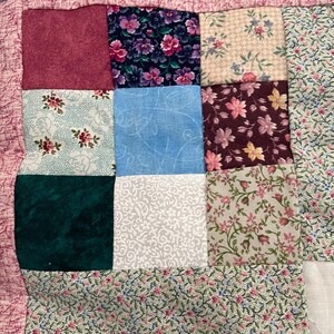 May include: A patchwork quilt featuring a variety of square fabric blocks in diverse patterns and colors, including floral prints in pink, green, and purple, solid dark teal, textured light blue, and white with a delicate vine pattern.