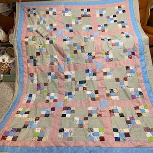 May include: A patchwork quilt with a gray background and a pink and blue border. The quilt is made up of many small squares of different fabrics, each with a different pattern. The quilt is folded in half and is hanging on a clothesline.