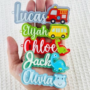 May include: A collection of colorful name tags with various designs. Each tag features a name in a unique font and color, accompanied by a related cartoon image, such as a fire truck, school bus, or dinosaur. The names include Lucas, Elijah, Chloe, Jack, and Olivia.