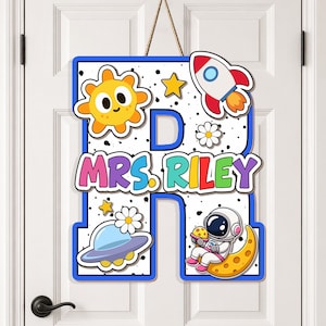 May include: A wooden door sign in the shape of the letter "D" with the text "MRS. RILEY". The sign features space-themed illustrations, including a sun, rocket, astronaut, and UFO, with a blue border and a rope for hanging.