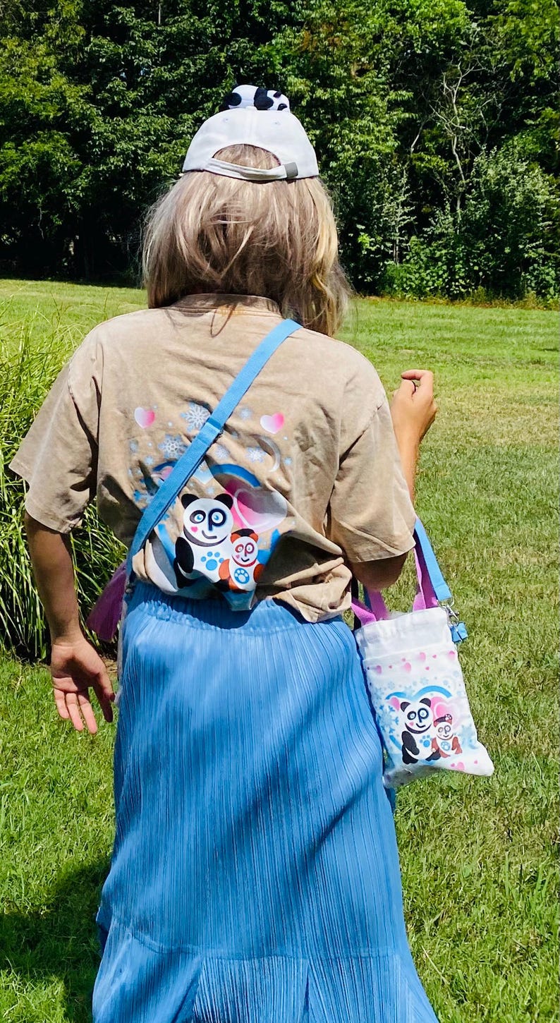Puede incluir: Una camiseta color beige con un gr&aacute;fico de oso panda, falda plisada azul y gorra de b&eacute;isbol blanca con un dise&ntilde;o de panda. Un peque&ntilde;o bolso tote blanco con un gr&aacute;fico de oso panda se lleva al hombro con una correa azul.