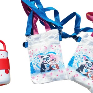 Puede incluir: Bolsos cruzados de lona blanca con un dise&ntilde;o de oso panda. Los bolsos tienen correas azules y rosas. El dise&ntilde;o del panda incluye un coraz&oacute;n, copos de nieve y un &aacute;rbol de Navidad. Tambi&eacute;n se ve una botella de agua roja y blanca.