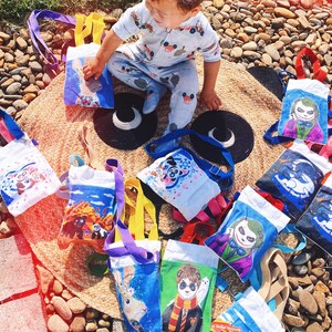 Puede incluir: Una colecci&oacute;n de peque&ntilde;as bolsas de mano con varios dise&ntilde;os de personajes de dibujos animados, incluyendo un panda, un conejo y el Joker. Las bolsas tienen correas de diferentes colores y est&aacute;n dispuestas sobre una estera tejida. Un ni&ntilde;o con un mono azul claro est&aacute; sentado entre las bolsas.
