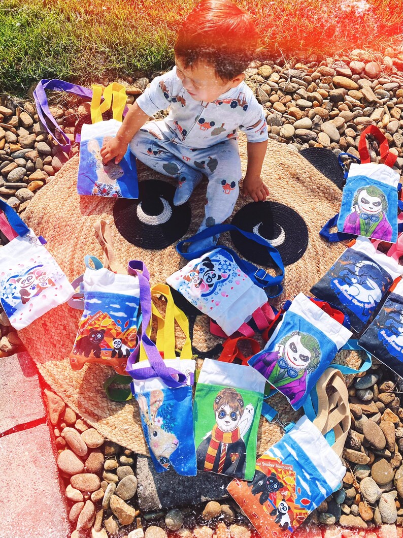 Puede incluir: Una colecci&oacute;n de peque&ntilde;as bolsas de mano con varios dise&ntilde;os de personajes de dibujos animados, incluyendo un panda, un conejo y el Joker. Las bolsas tienen correas de diferentes colores y est&aacute;n dispuestas sobre una estera tejida. Un ni&ntilde;o con un mono azul claro est&aacute; sentado entre las bolsas.