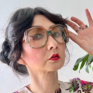 May include: Oversized brown and green eyeglasses with a retro design. The glasses have a thick frame and tinted lenses. The person is wearing a colorful paisley print top with shades of pink, green, and purple.