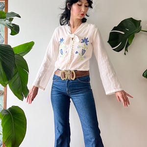 May include: A white long-sleeved blouse with a tie-neck, featuring blue and green floral embroidery on the chest, paired with dark blue denim flare jeans, a brown leather belt with a gold buckle, and red platform sandals with cut-out details.