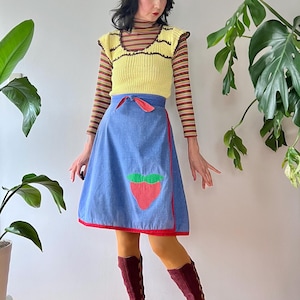 May include: A vintage outfit featuring a yellow knit top with brown trim over a striped long-sleeve shirt, paired with a blue denim skirt with a red trim and a strawberry applique. The outfit is completed with burgundy knee-high boots.