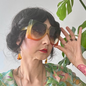 May include: Oversized square sunglasses with a gradient lens, transitioning from green to orange. The sunglasses are set against a backdrop of a floral patterned top and a green plant. The frame is a light color.