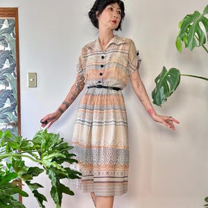 Könnte beinhalten: Ein Vintage-Midikleid mit Kragen und kurzen Ärmeln. Das Kleid hat eine Knopfleiste vorne, einen schwarzen Gürtel und ein gemustertes Design in Pfirsich-, Creme- und Blautönen. Das Kleid wird mit bordeauxfarbenen Sandalen mit Blockabsatz getragen.