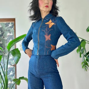 Puede incluir: Conjunto de chaqueta y pantalón vaquero. La chaqueta tiene cremallera frontal y bordados de mariposas en tonos naranja y marrón. El pantalón es de pernera ancha. El conjunto es de estilo vintage.